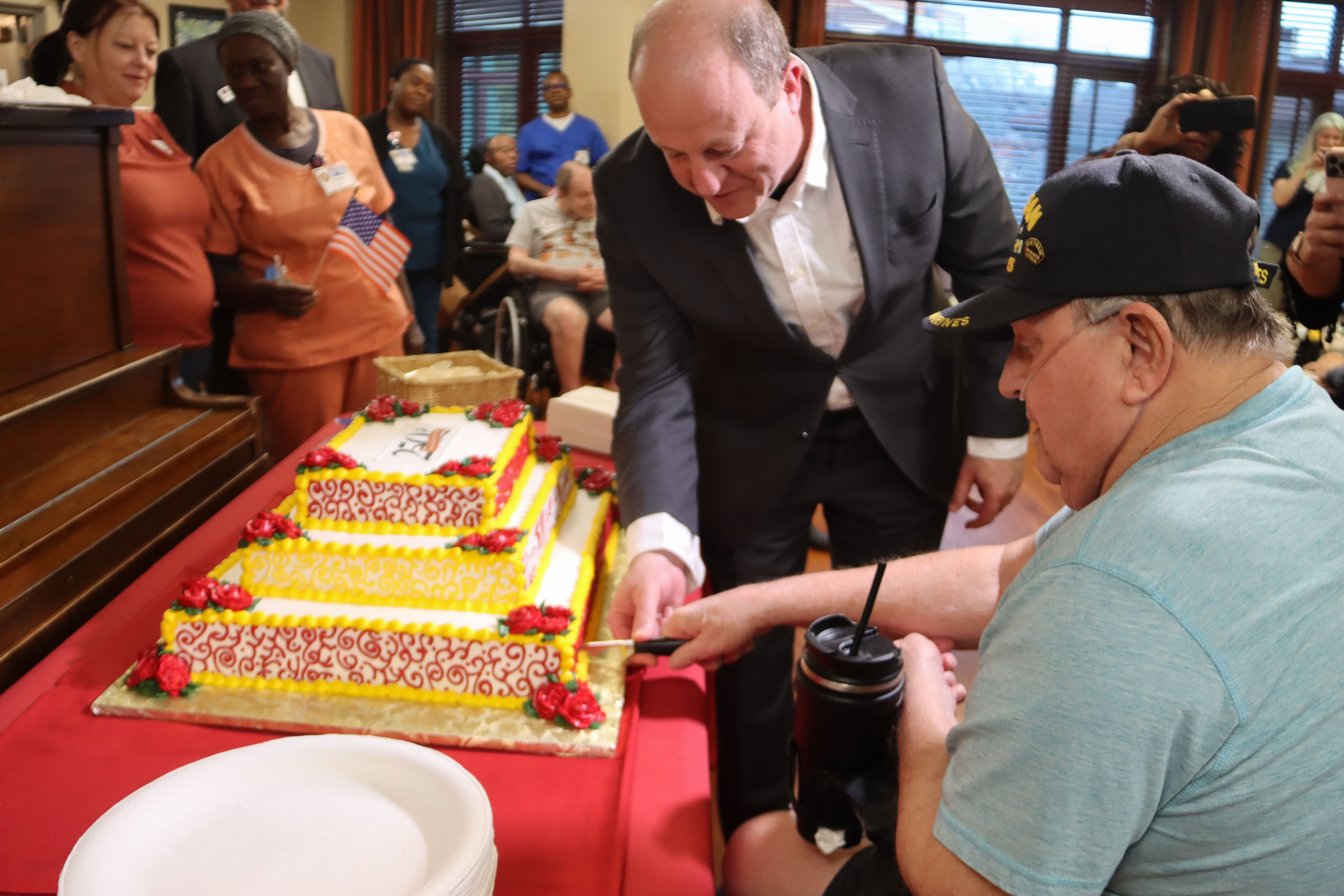 Governor Polis cuts a cake for the Marine Corps' 250th birthday alongside a veteran at the Veterans Community Living Center at Fitzsimons. 